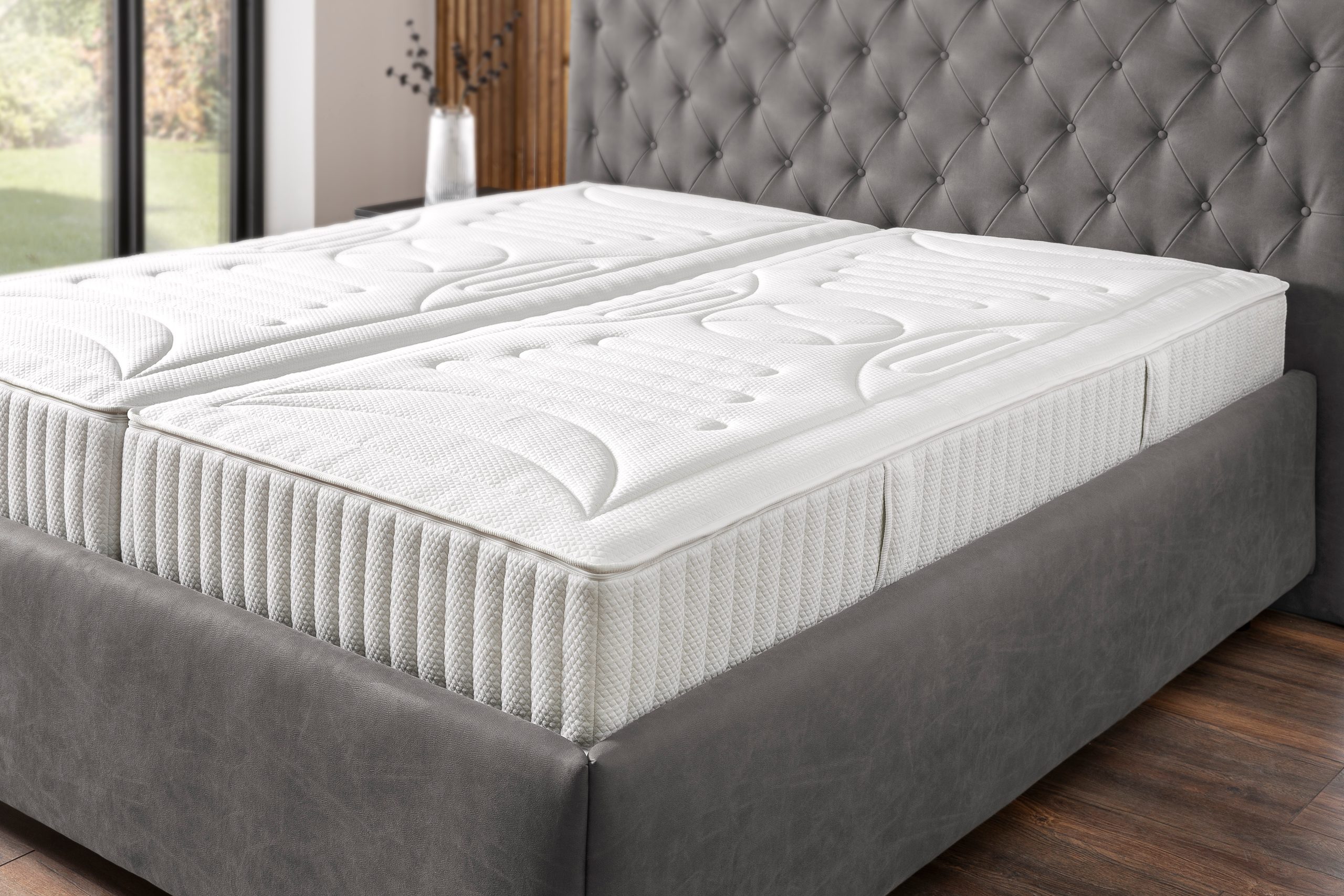 Zwei unbezogene Matratzen liegen nebeneinander auf einem grauen Boxspringrahmen: die gesteppte Oberseite, die genähte Kante und die profilierte Seitenwange sind offen sichtbar.
